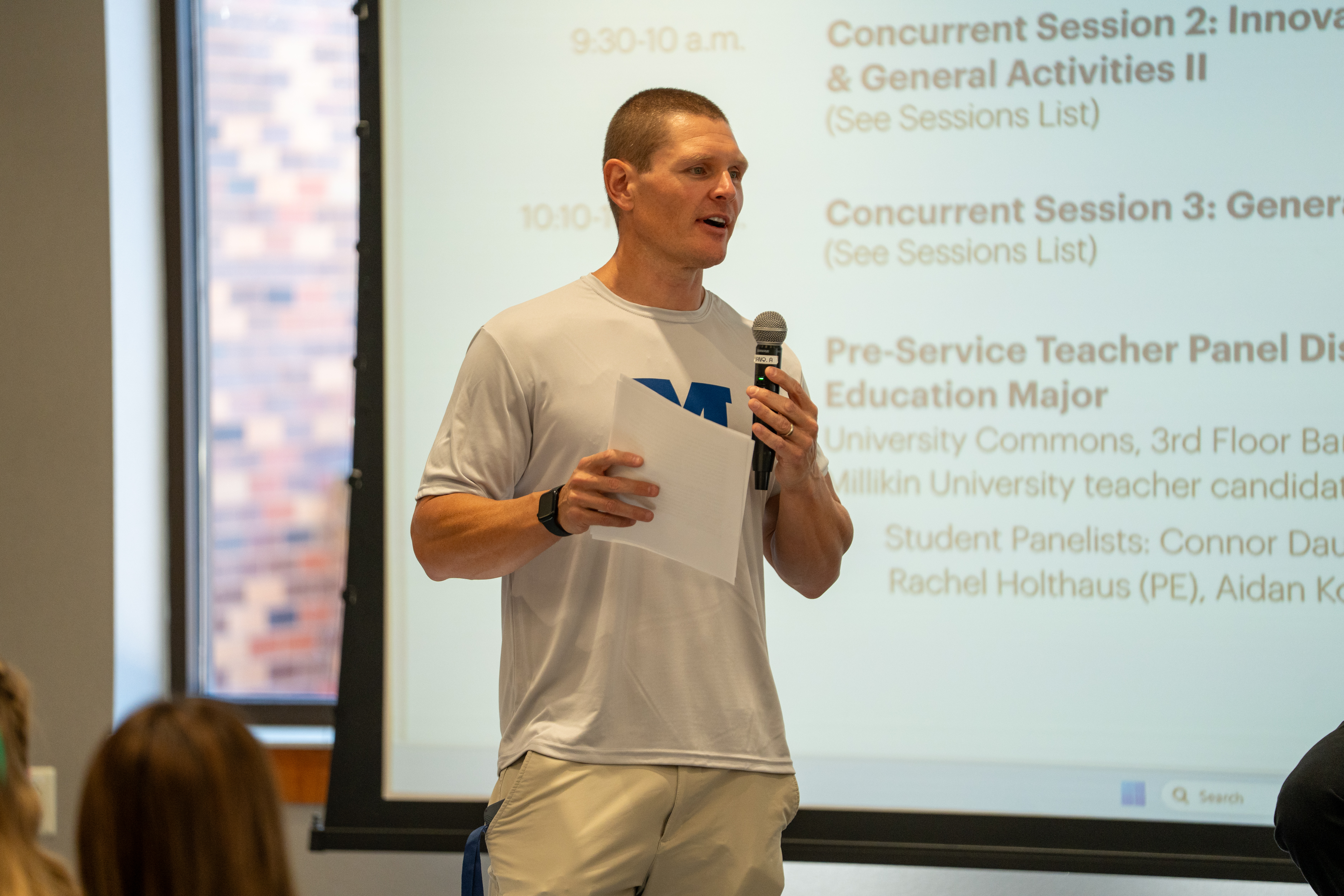 A speaker stands in front of a projected conference schedule, holding a microphone and a sheet of paper while addressing an audience. The speaker wears a white T-shirt with a blue “M” logo and light-colored pants. Visible text on the screen behind includes references to concurrent sessions and a pre-service teacher panel discussion at Millikin University.