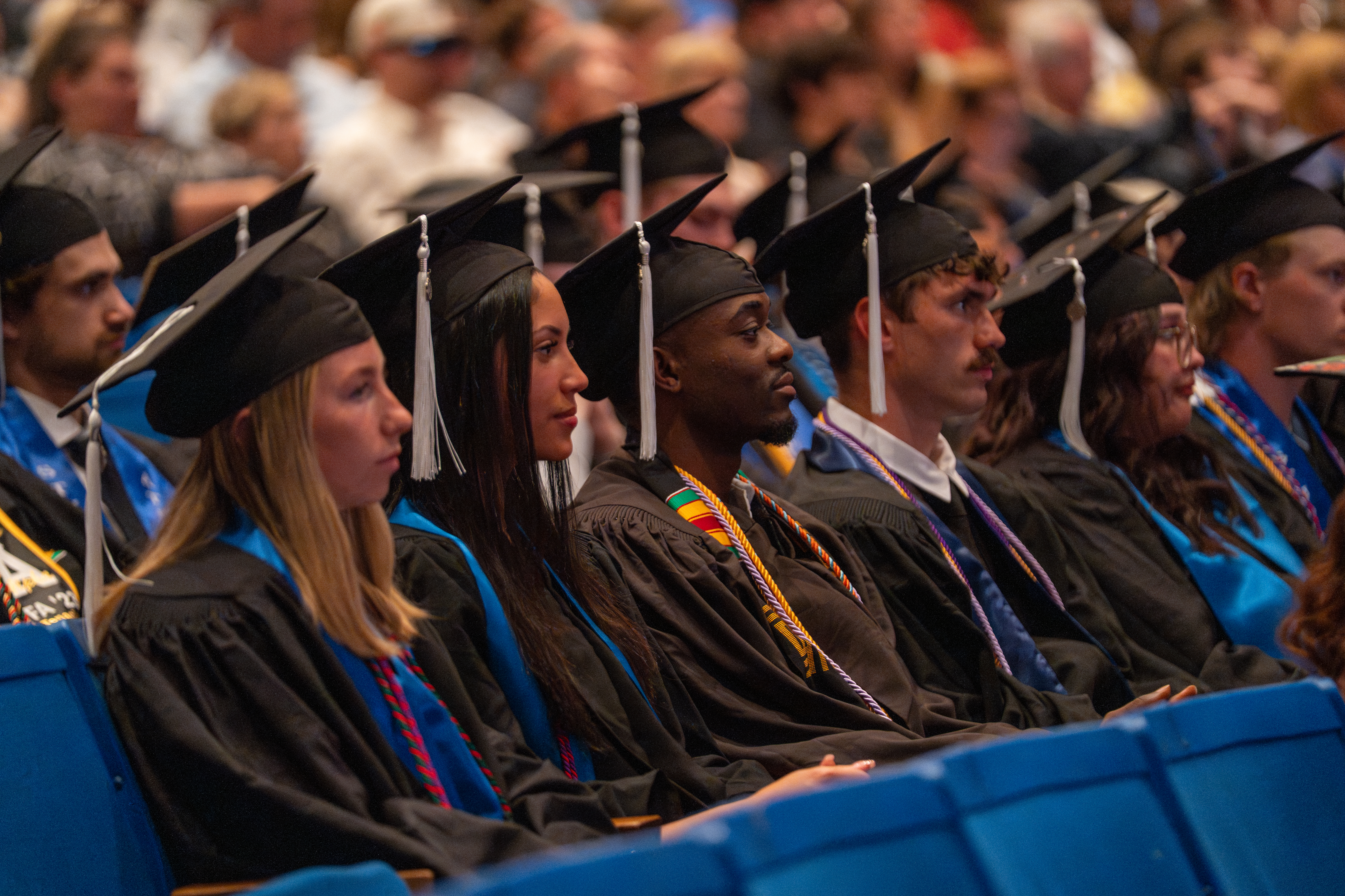 Graduates in black caps and gowns sit in rows during a commencement ceremony, listening attentively as they await the conferral of their degrees. Several wear honor cords and stoles, with a blurred audience visible in the background.