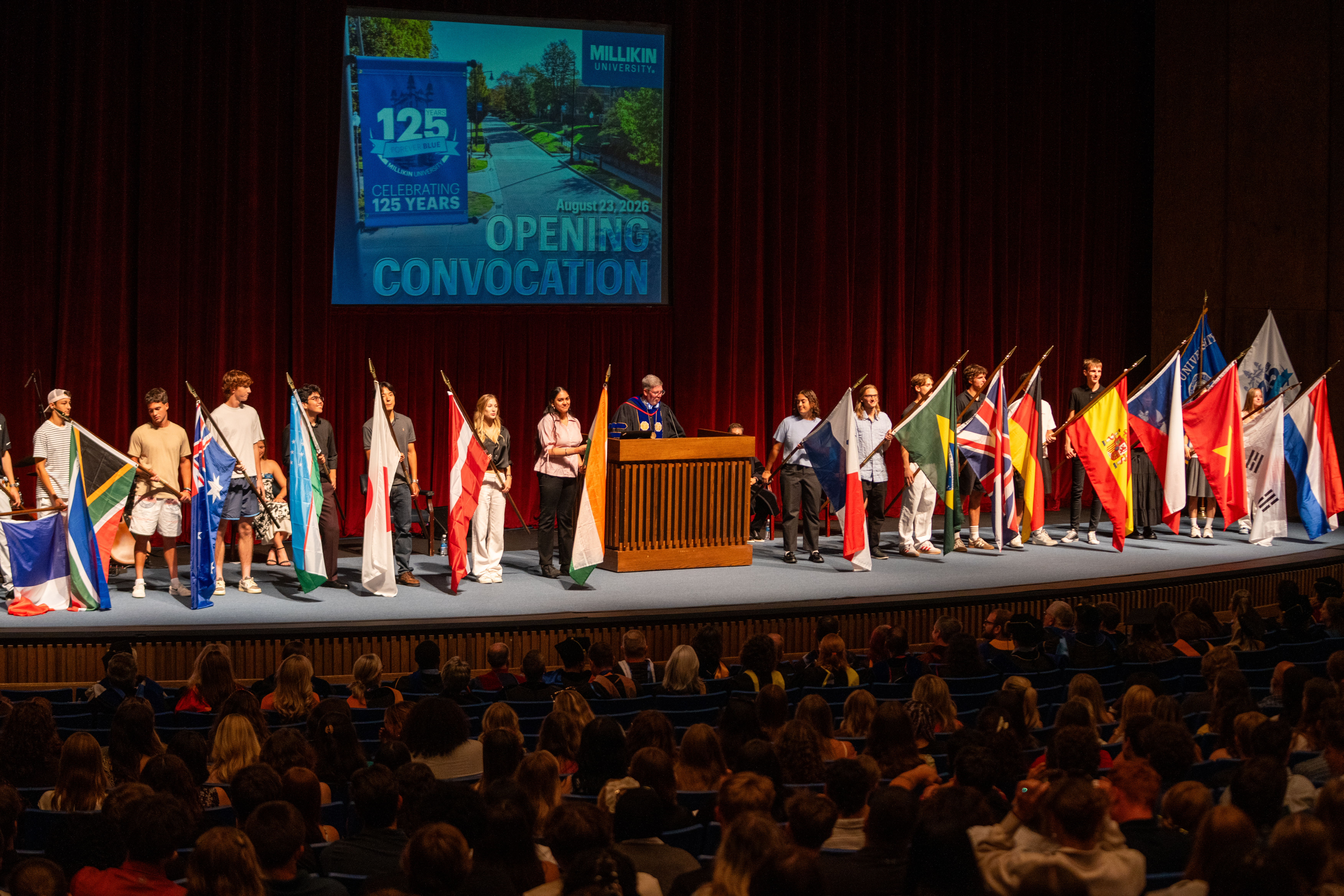 Students carrying flags from countries around the world stand across a theater stage during Millikin University’s Opening Convocation. A speaker stands at a podium in the center, while an audience fills the auditorium. A large screen above the stage displays “Opening Convocation,” “August 23, 2026,” and Millikin University’s 125th anniversary celebration.
