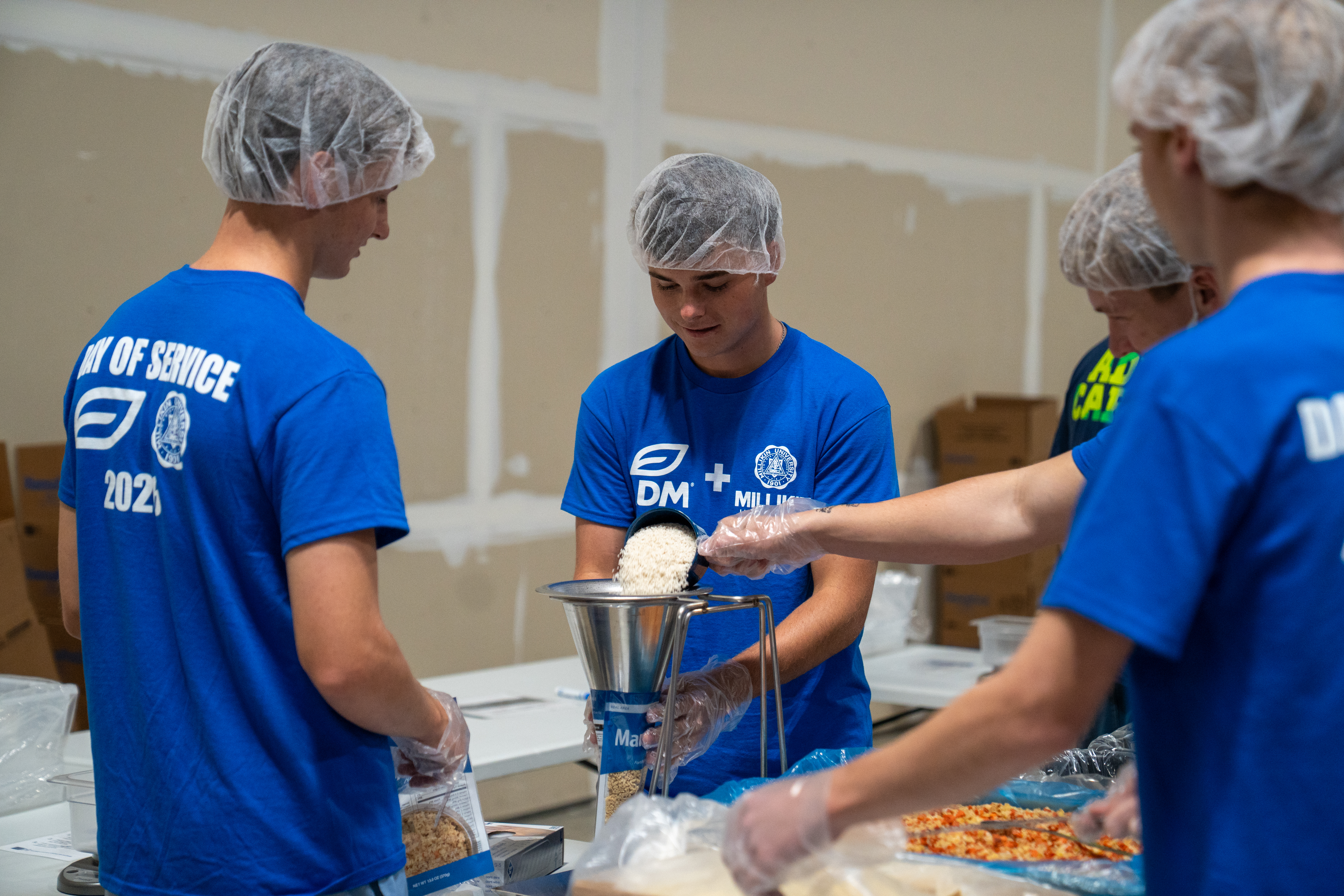 Volunteers wearing blue ADM-Millikin Day of Service T-shirts, hairnets, and gloves work at a meal-packing station inside a warehouse. One volunteer pours rice through a metal funnel into a food package while others add ingredients from large containers at the table. Visible shirt text includes “ADM + Millikin University” and “Day of Service 2026,” with boxes and supplies arranged around the packing area.