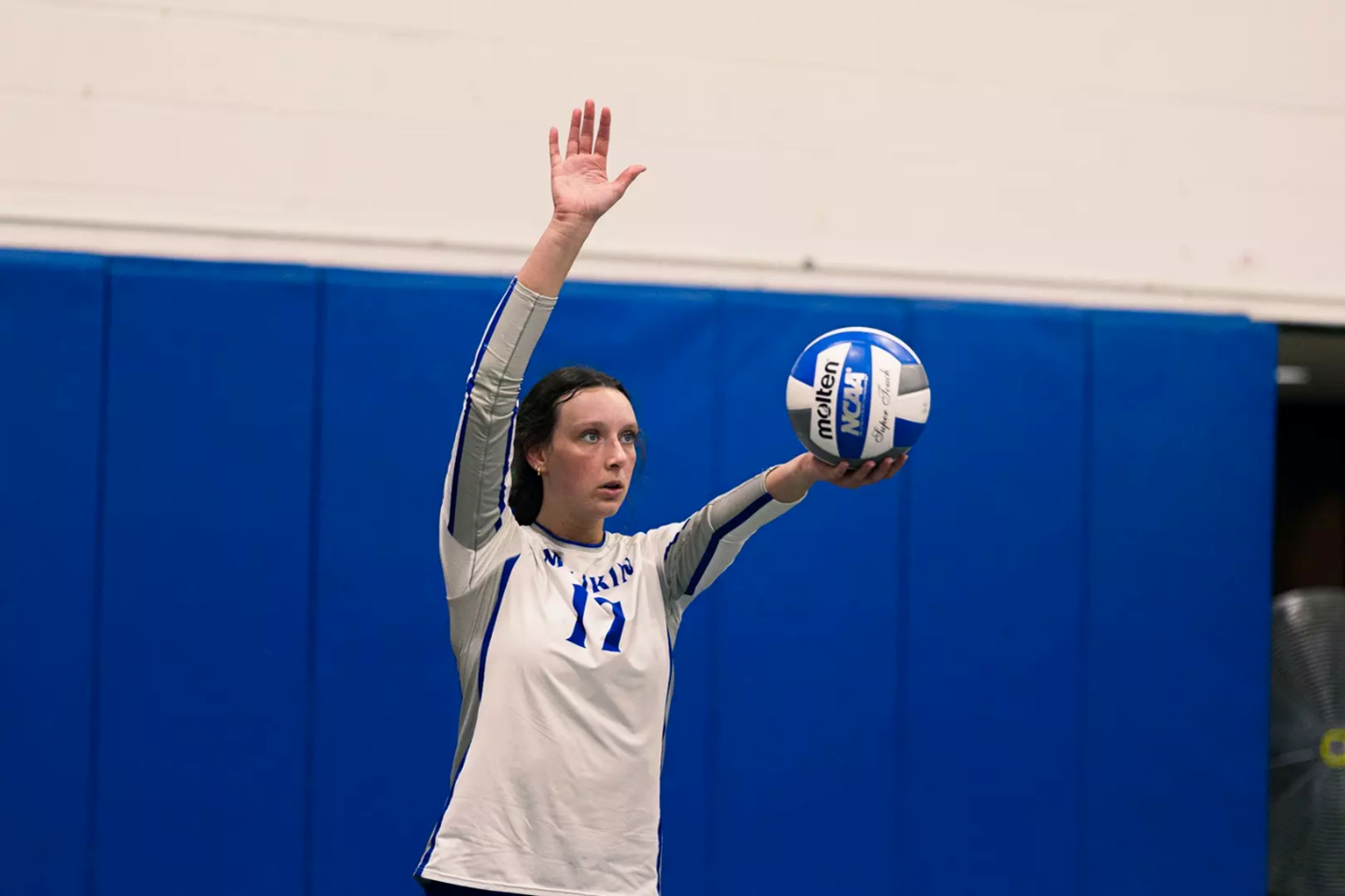 Volleyball player wearing a white jersey with the number 17 prepares to serve during an indoor match, holding a blue-and-white volleyball in one hand while raising the other arm overhead in front of a blue padded gym wall.