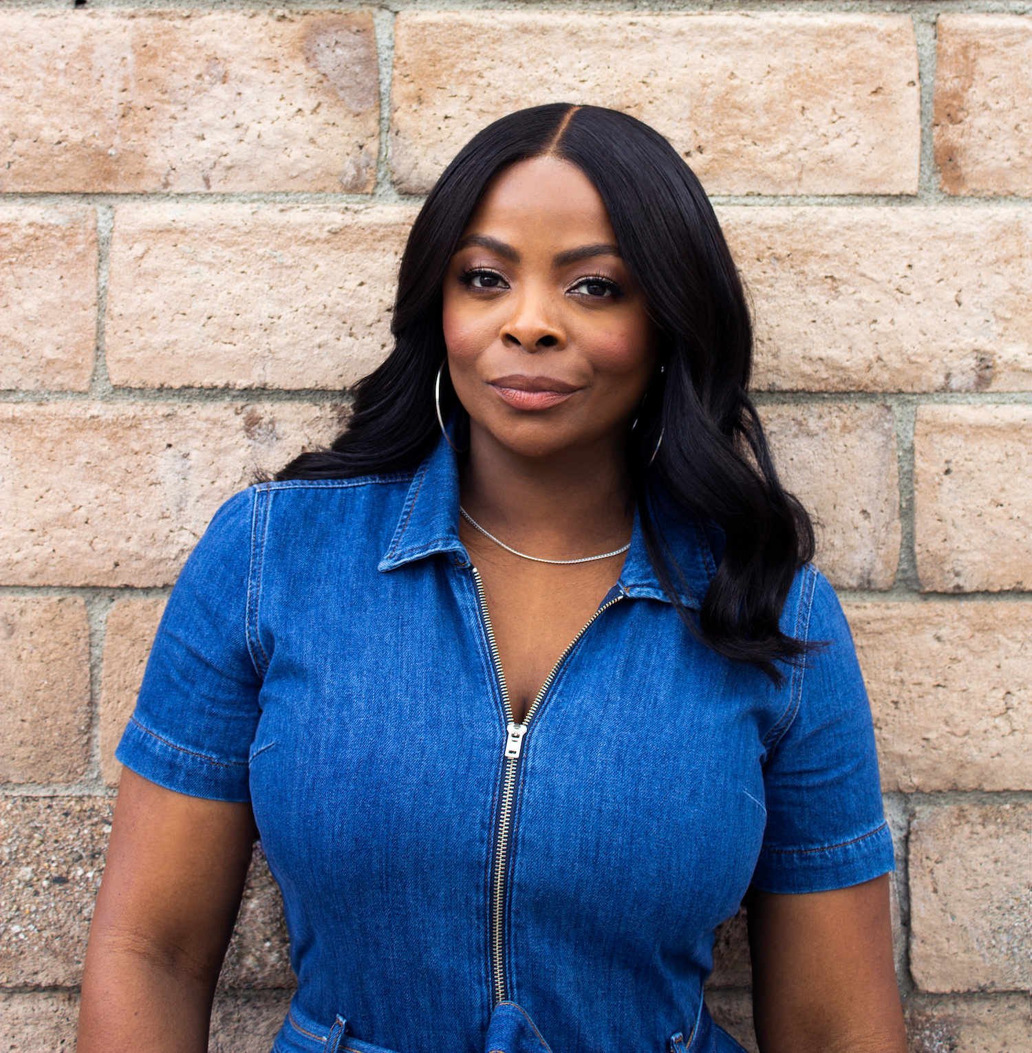 Janelle James, comedian, stands in front of a brick wall. She is wearing a blue jumpsuit and silver hoop earrings.