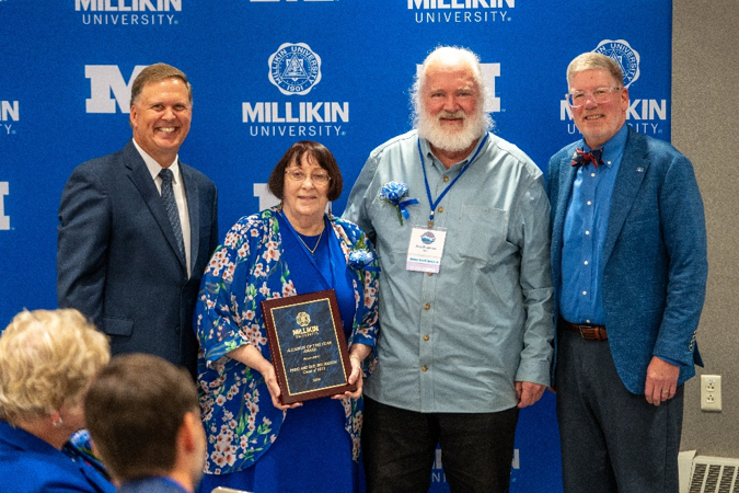 Four individuals pose for a photo in front of a Millikin University backdrop, with one person holding a commemorative plaque during an awards recognition event.