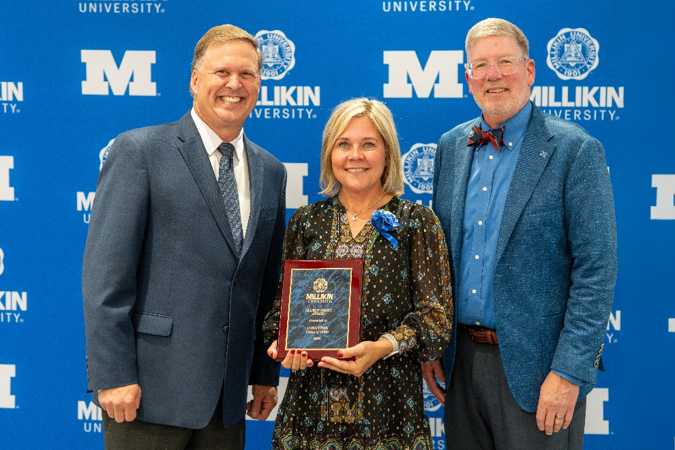 Three individuals pose in front of a Millikin University backdrop, with the person in the center holding an alumni award plaque during a recognition ceremony.