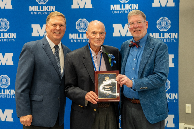 Three individuals pose in front of a Millikin University backdrop, with the person in the center holding a distinguished alumni award plaque during a recognition ceremony.