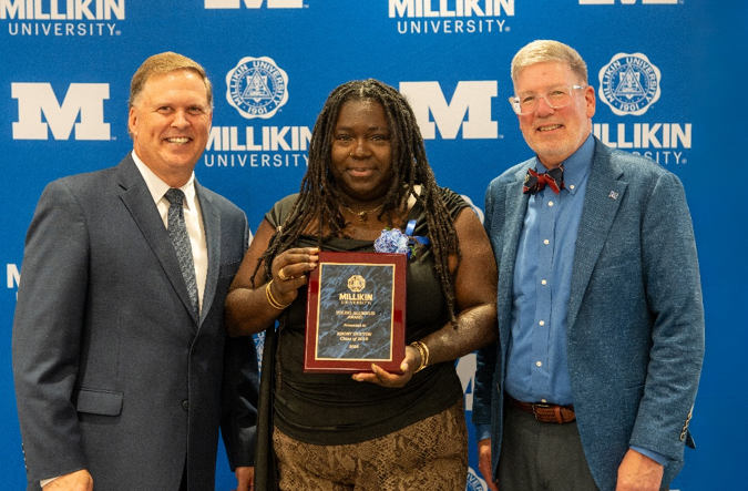 Three individuals pose in front of a Millikin University backdrop, with the person in the center holding a Young Alumni Award plaque during a recognition ceremony.