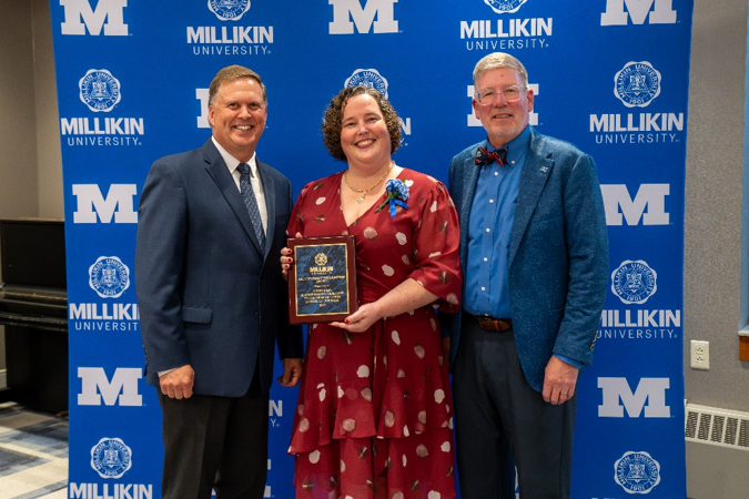 Three individuals pose in front of a Millikin University backdrop, with the person in the center holding an alumni award plaque during a recognition ceremony.