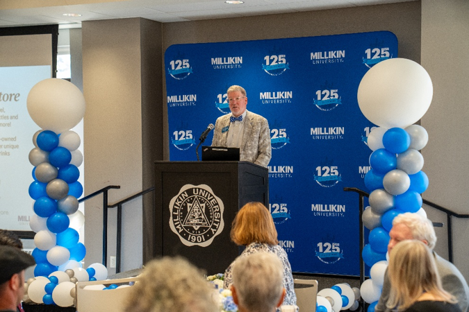 A speaker stands at a podium bearing the Millikin University seal, addressing attendees seated at banquet tables. The stage is framed by blue, white, and silver balloon columns and a blue backdrop featuring Millikin University’s 125th anniversary branding. A projection screen is visible to the left, and the event takes place in a well-lit indoor gathering space decorated for a university recognition or celebration program.