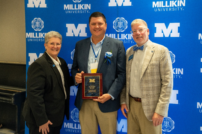 Three attendees stand together for a recognition photo at a Millikin University event. The individual in the center displays an award plaque while wearing a blue-and-white boutonniere and event ribbon. The group is posed before a bright blue backdrop featuring Millikin University branding and institutional seals, with a piano visible along the edge of the room.
