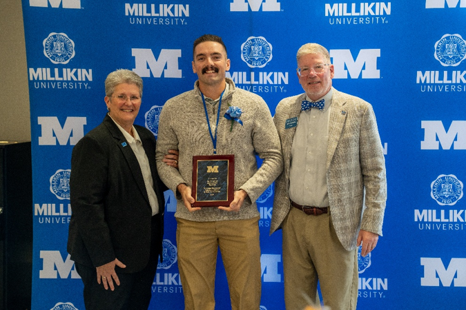 An award recipient stands between two university representatives while holding a maroon-and-gold recognition plaque at a Millikin University event. The recipient wears a blue ribbon and boutonniere, and the group poses in front of a blue backdrop featuring Millikin University logos and large white “M” graphics. The formal portrait highlights the presentation of the award during a campus recognition ceremony.