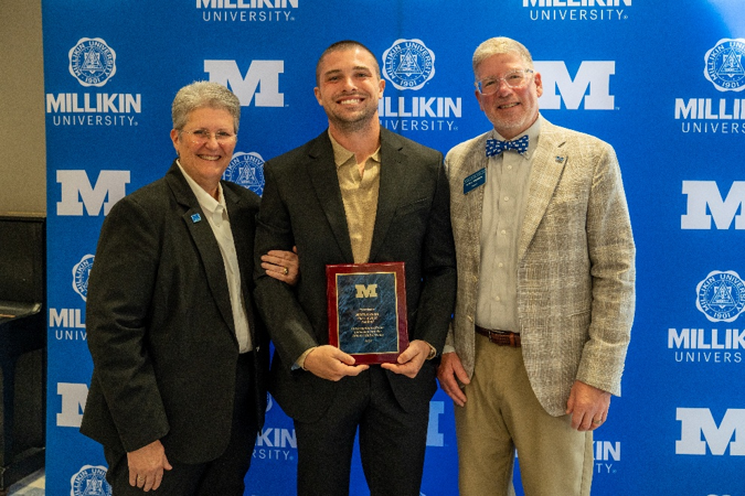 A plaque recipient poses with two university representatives during a Millikin University recognition event. Standing before a blue backdrop covered with Millikin logos, the recipient holds a maroon-and-gold award plaque while wearing a dark suit jacket and light-colored shirt. The image captures a formal awards presentation photo, with the plaque prominently displayed at the center of the group.