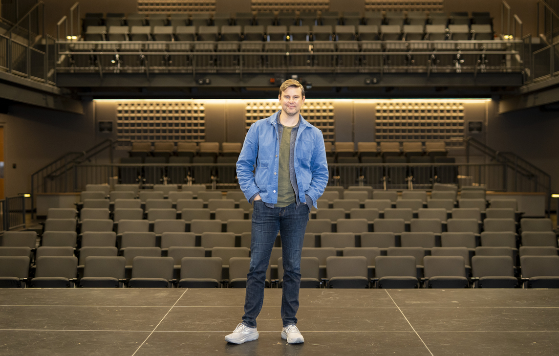 A person stands at the center of a theatre stage facing the camera, wearing a blue jacket, dark jeans, and light-colored shoes. Rows of empty auditorium seats fill the foreground and middle ground, while tiered seating and balconies rise in the background. Warm overhead lighting illuminates the modern performance venue, highlighting the symmetry of the stage and audience seating.