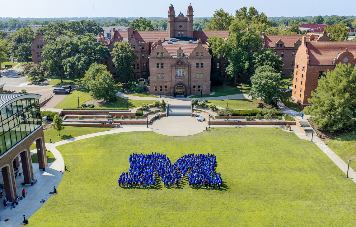 Aerial view of a college campus courtyard on a sunny day. Hundreds of students wearing blue shirts stand on a large green lawn arranged in the shape of a capital “M,” with historic red-brick academic buildings, trees, walkways, and landscaped terraces in the background.
