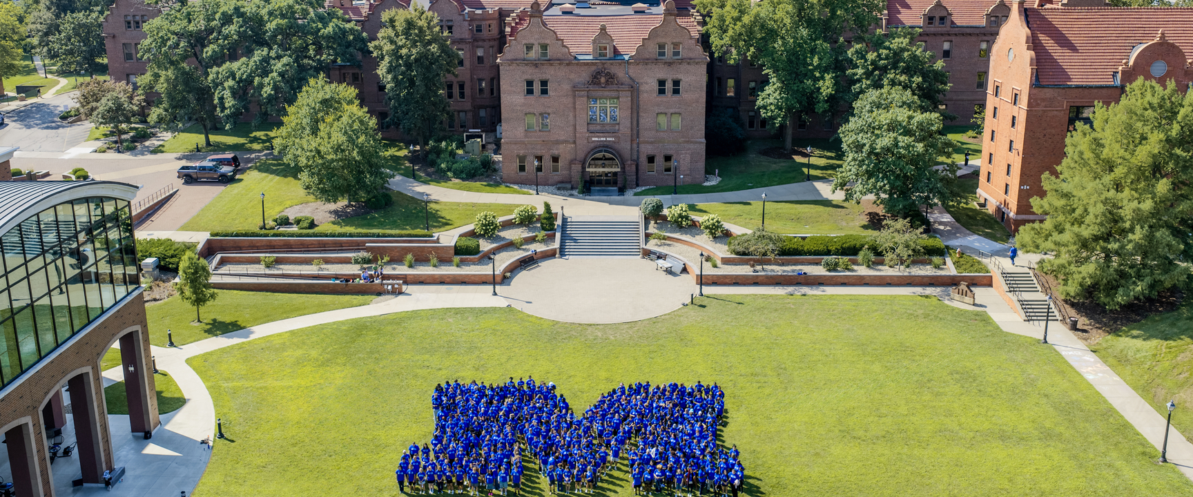 Aerial view of a college campus courtyard on a sunny day. Hundreds of students wearing blue shirts stand on a large green lawn arranged in the shape of a capital “M,” with historic red-brick academic buildings, trees, walkways, and landscaped terraces in the background.