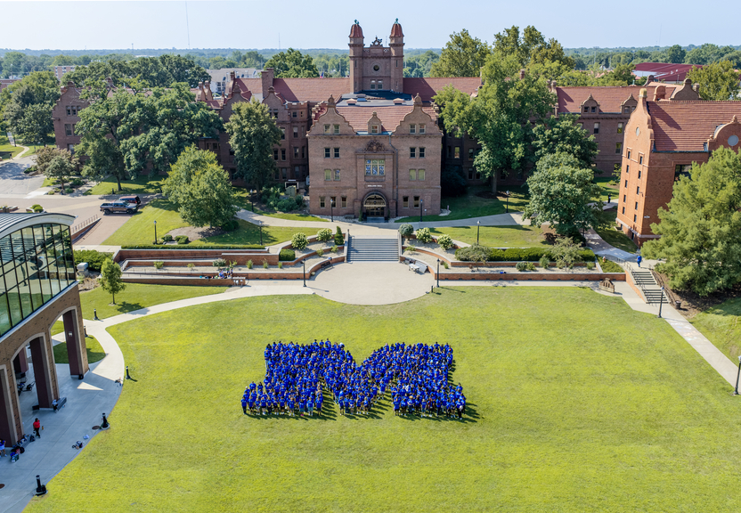 Aerial view of a college campus courtyard on a sunny day. Hundreds of students wearing blue shirts stand on a large green lawn arranged in the shape of a capital “M,” with historic red-brick academic buildings, trees, walkways, and landscaped terraces in the background.