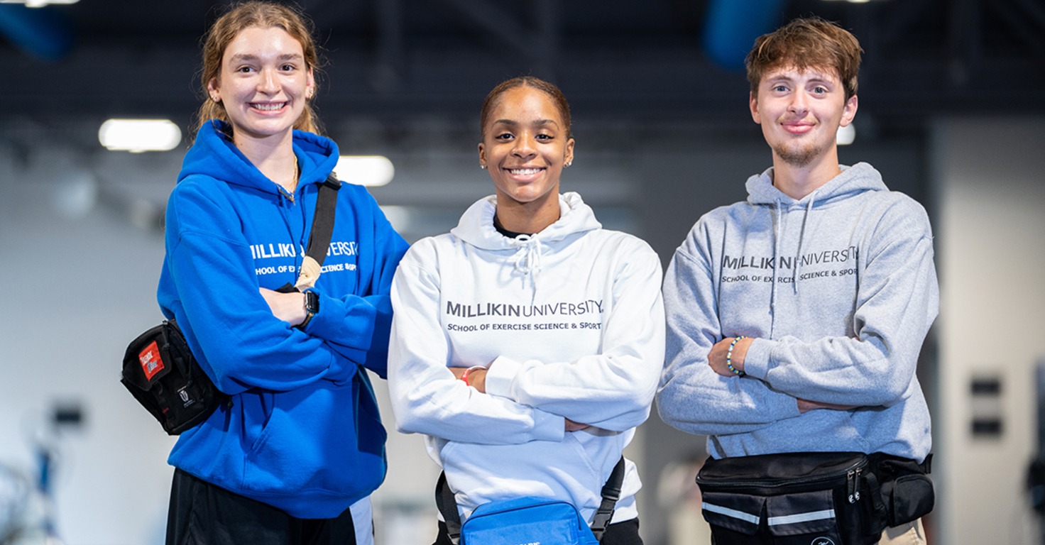 Three Millikin University students stand side by side in the Exercise Science & Sport Facility, smiling at the camera with their arms crossed. They are wearing Millikin hooded sweatshirts in blue, white, and gray, along with waist packs. Exercise equipment is visible in the softly blurred background.