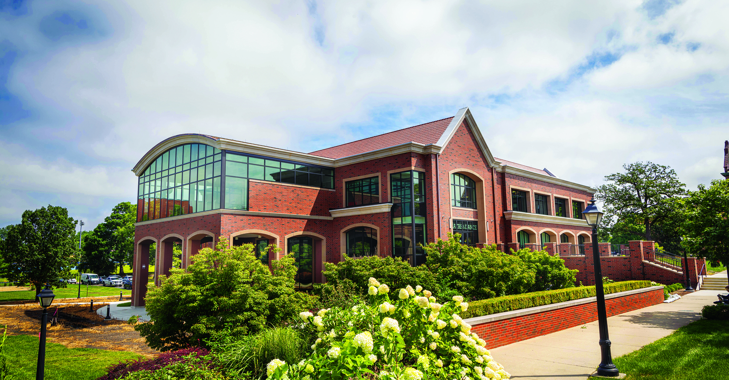 Brick academic building with large glass windows, landscaped shrubs, and white flowers in the foreground, under a partly cloudy sky.