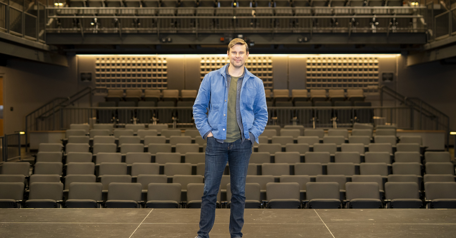 A person stands at the center of a theatre stage facing the camera, wearing a blue jacket, dark jeans, and light-colored shoes. Rows of empty auditorium seats fill the foreground and middle ground, while tiered seating and balconies rise in the background. Warm overhead lighting illuminates the modern performance venue, highlighting the symmetry of the stage and audience seating.