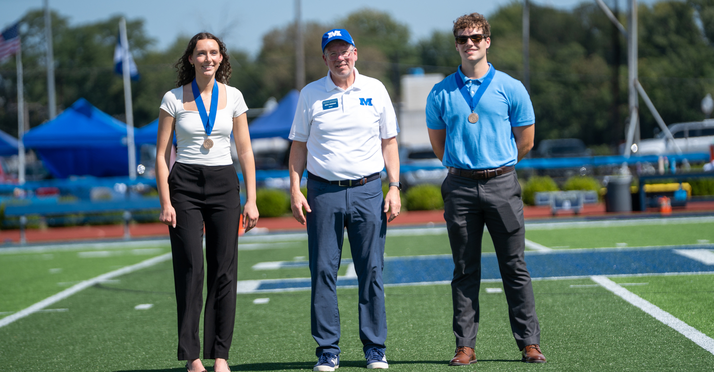 Three individuals wearing medals stand on a football field during an outdoor ceremony. The person in the center wears a white polo shirt and blue cap with an “M” logo, while the two individuals on either side wear business-casual attire. Blue tents, field equipment, and trees are visible in the background under a clear sky.