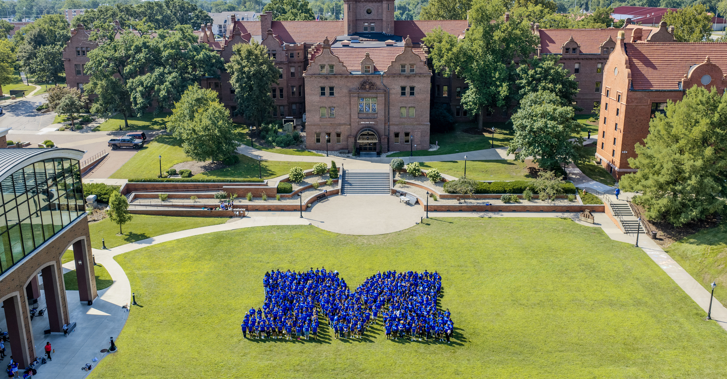 Aerial view of a college campus courtyard on a sunny day. Hundreds of students wearing blue shirts stand on a large green lawn arranged in the shape of a capital “M,” with historic red-brick academic buildings, trees, walkways, and landscaped terraces in the background.