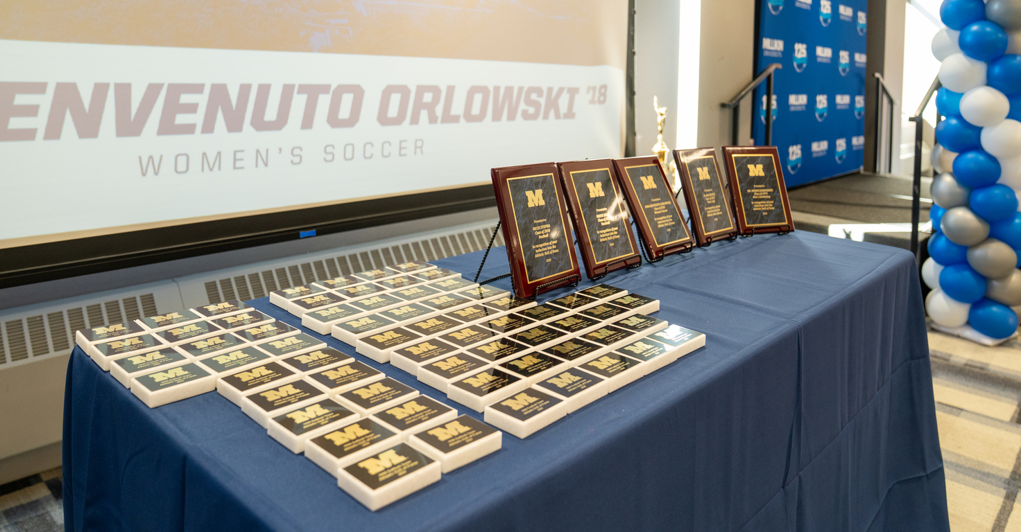 A display table covered with a blue tablecloth holds dozens of black-and-gold awards and five larger framed plaques arranged in rows. Behind the table, a large projection screen displays “Benvenuto Orlowski ’18 Women’s Soccer.” A blue-and-white balloon column and a branded backdrop stand to the right, suggesting an athletic awards or Hall of Fame recognition event.