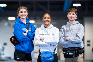 Three Millikin University students stand side by side in the Exercise Science & Sport Facility, smiling at the camera with their arms crossed. They are wearing Millikin hooded sweatshirts in blue, white, and gray, along with waist packs. Exercise equipment is visible in the softly blurred background.