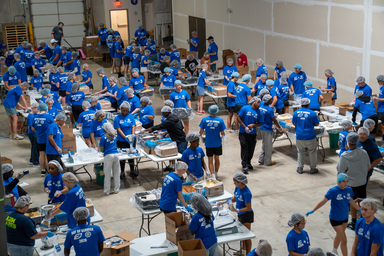 Large indoor warehouse space filled with dozens of volunteers wearing matching blue T-shirts and hairnets, working at long tables to assemble and package meals. Boxes, trays, and food-packaging materials are spread across the tables, while volunteers move between stations in an organized assembly-line setup. The high-ceiling room has unfinished walls, industrial lighting, and stacks of supplies visible in the background.