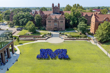 Aerial view of a college campus courtyard on a sunny day. Hundreds of students wearing blue shirts stand on a large green lawn arranged in the shape of a capital “M,” with historic red-brick academic buildings, trees, walkways, and landscaped terraces in the background.