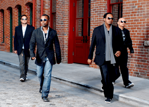 The band members of All-4-One walk down a street in front of a brick building.