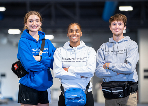Three Millikin University students stand side by side in the Exercise Science & Sport Facility, smiling at the camera with their arms crossed. They are wearing Millikin hooded sweatshirts in blue, white, and gray, along with waist packs. Exercise equipment is visible in the softly blurred background.