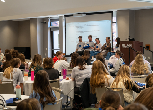Audience members seated at round tables listen to a panel discussion at a conference room. Five panelists sit at the front near a podium labeled “Millikin University,” with a presentation agenda projected on a screen behind them. Attendees face the speakers, and several water bottles and notebooks are visible on the tables throughout the room.