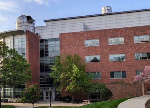 Modern multi-story red brick academic or office building with large glass windows and a central glass entrance. Mature green trees and landscaping frame the building, with a curved sidewalk leading through the foreground under a clear blue sky.