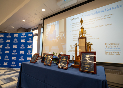 Awards plaques and a championship trophy displayed on a blue-draped table in a conference room, with a Millikin University backdrop and presentation screen in the background.